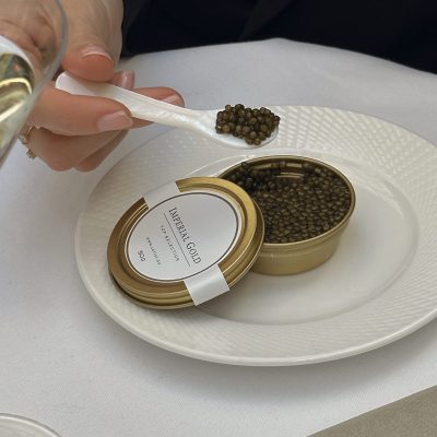 A person holding a spoonful of caviar above an open tin labeled "Imperial Gold, Top Selection." The caviar tin, containing 50 grams of high-quality caviar, is placed on a white textured plate. The person is using a white spoon, likely made of mother of pearl to ensure the delicate taste of the caviar is not compromised by metal utensils.